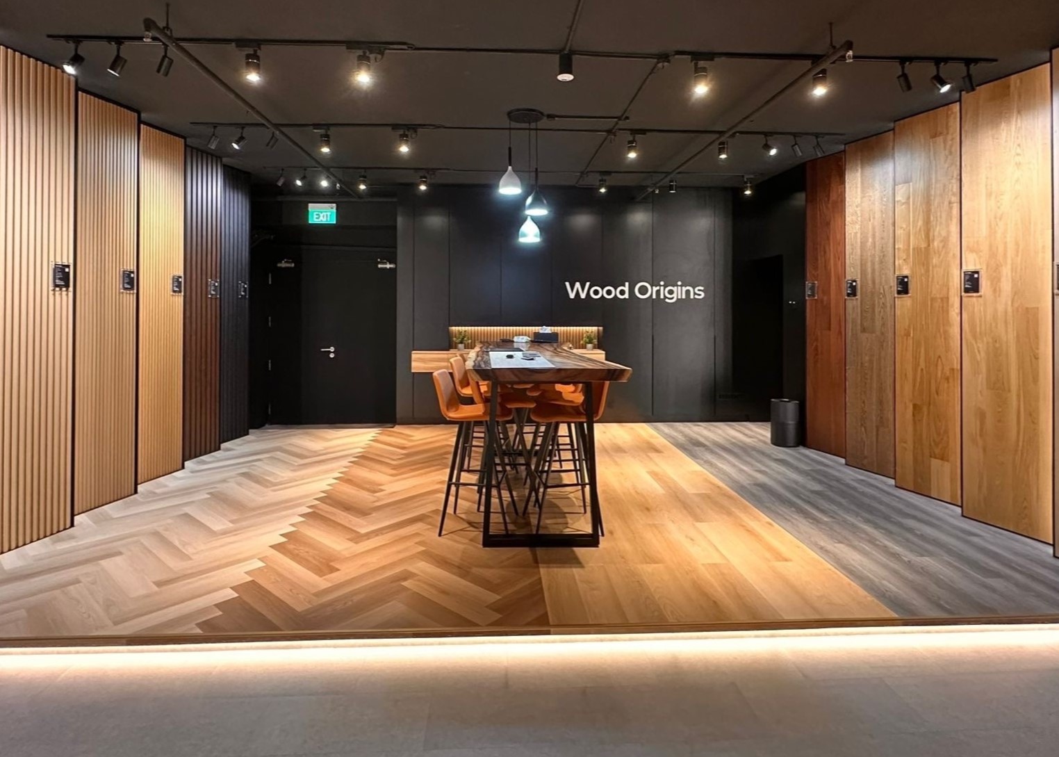 Wood Origins showroom in Singapore showcasing a variety of vinyl flooring designs, including herringbone and straight wood grain patterns. The space features a modern display with warm lighting, a central wooden table, and a stylish ambiance that highlights their premium flooring products.